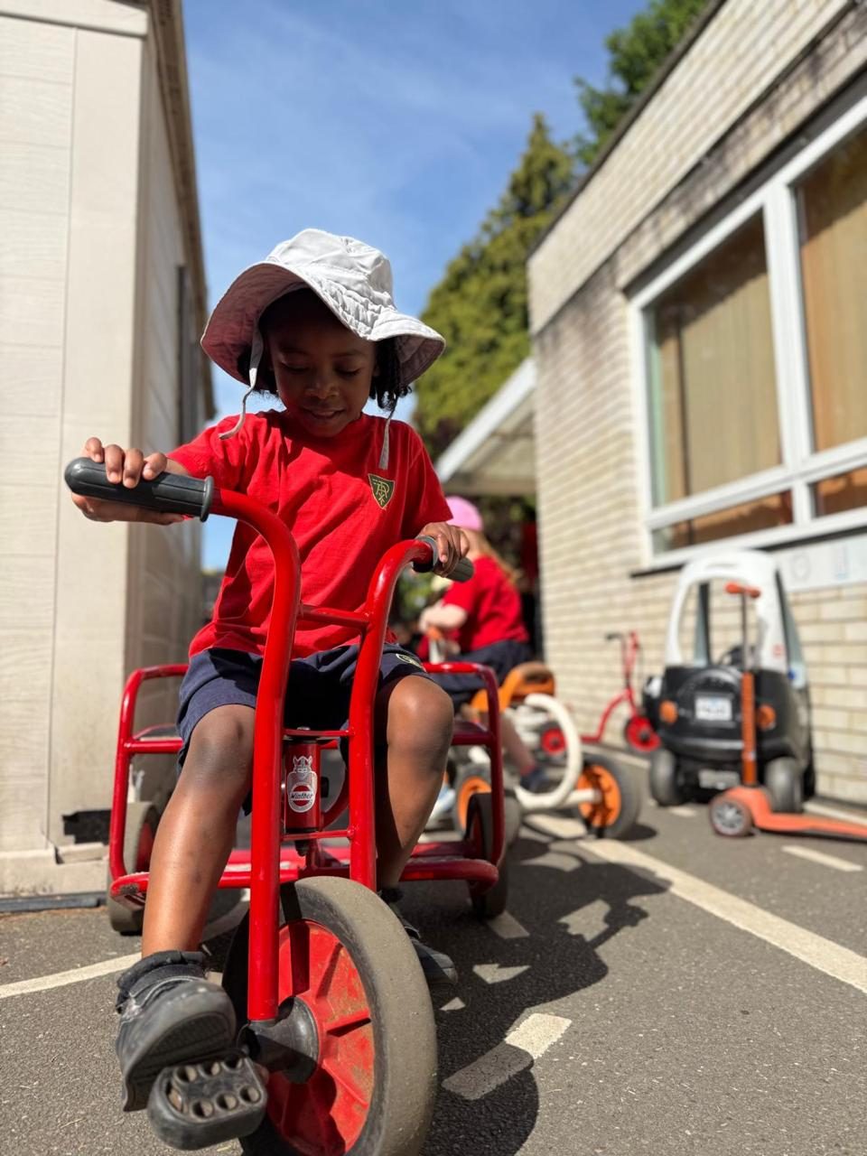 A nursery child pedals a trike outdoors on a sunny morning.