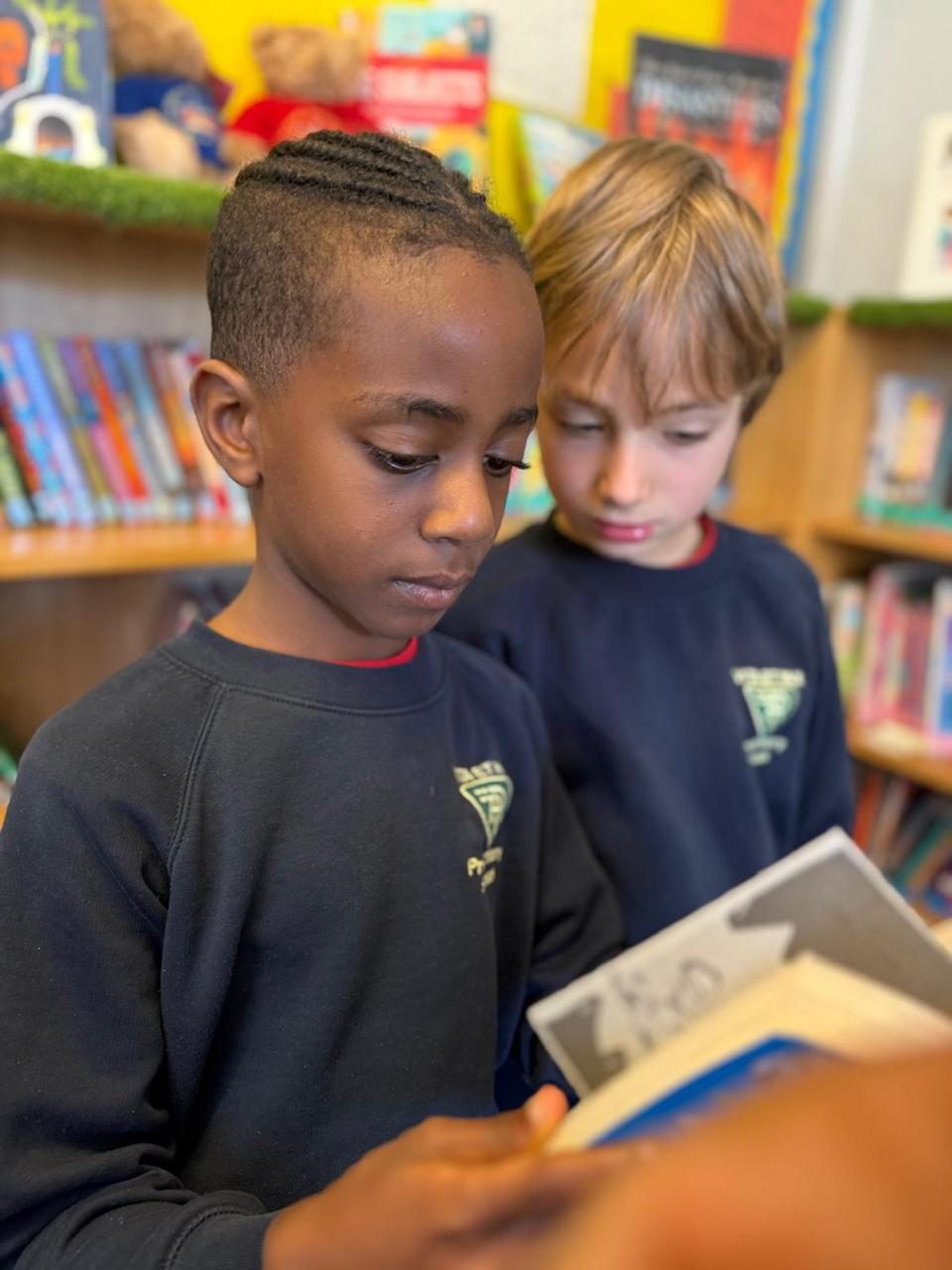 Two pupils read together among the library shelves.